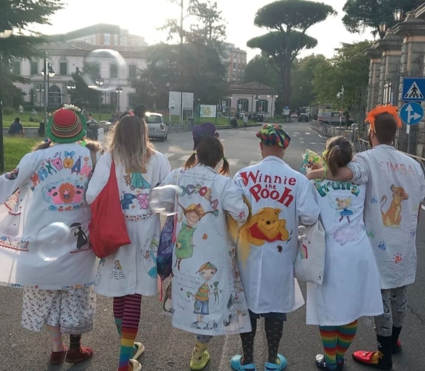 Volontari clown di Namo APS di spalle, con i camici dipinti a mano, camminano insieme verso l’ospedale